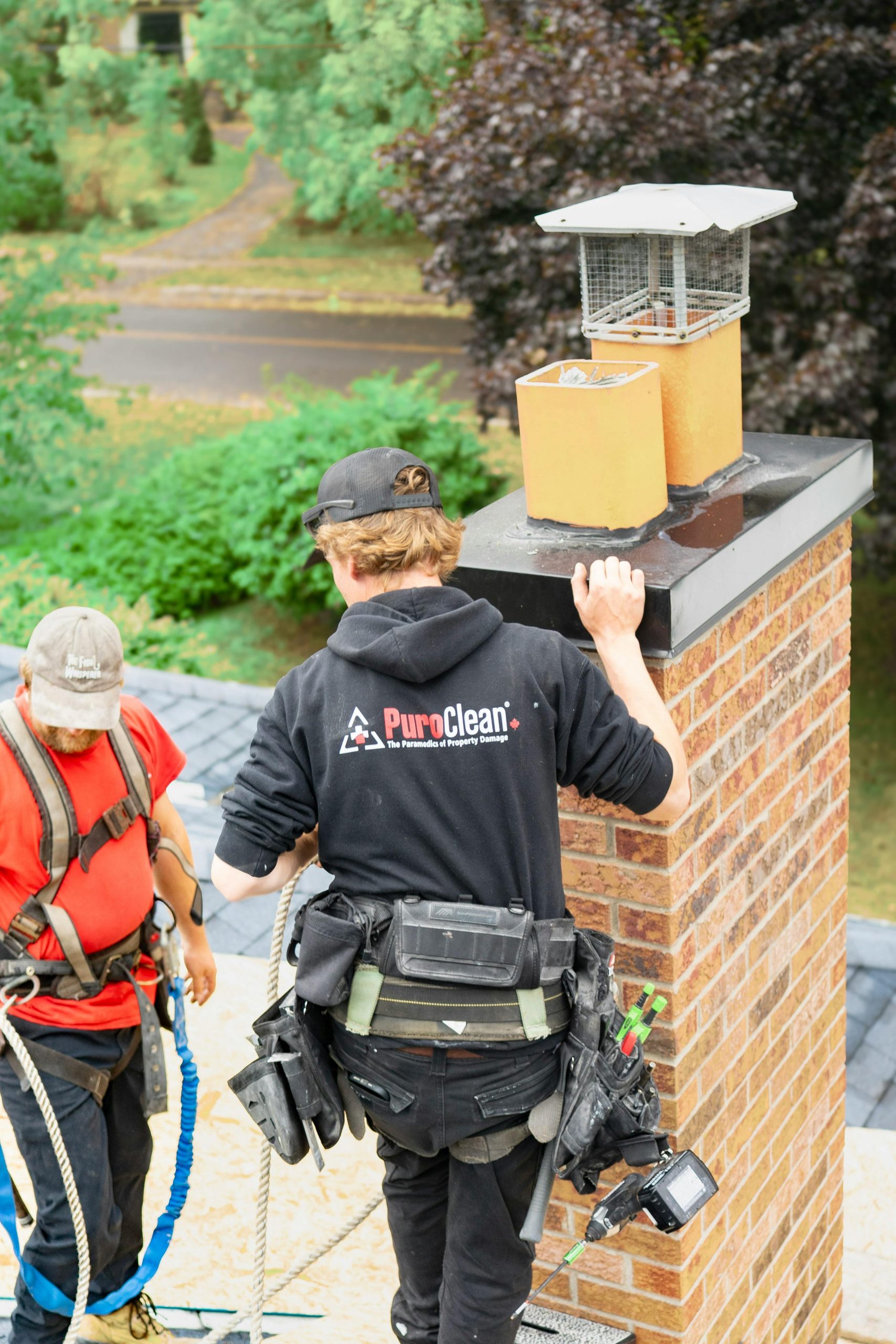 Two workers conducting a thorough chimney inspection on a rooftop in a wooded neighborhood. Deux ouvriers effectuant une inspection approfondie de cheminée sur un toit dans un quartier boisé.