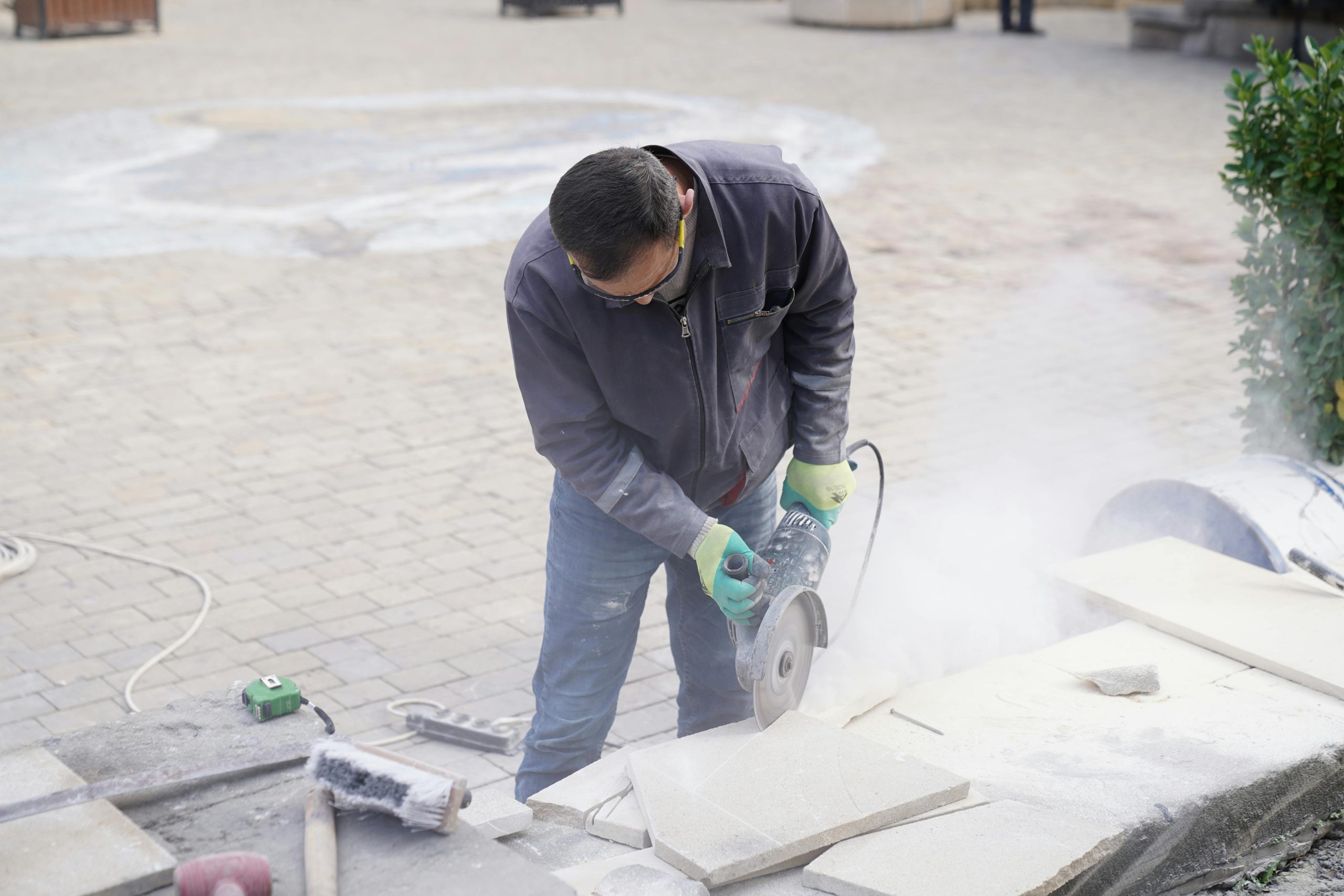 A construction worker uses a power saw to cut stone tiles outdoors, creating dust as he works. Un ouvrier du bâtiment utilise une scie électrique pour couper des carreaux de pierre à l'extérieur, créant de la poussière en travaillant.