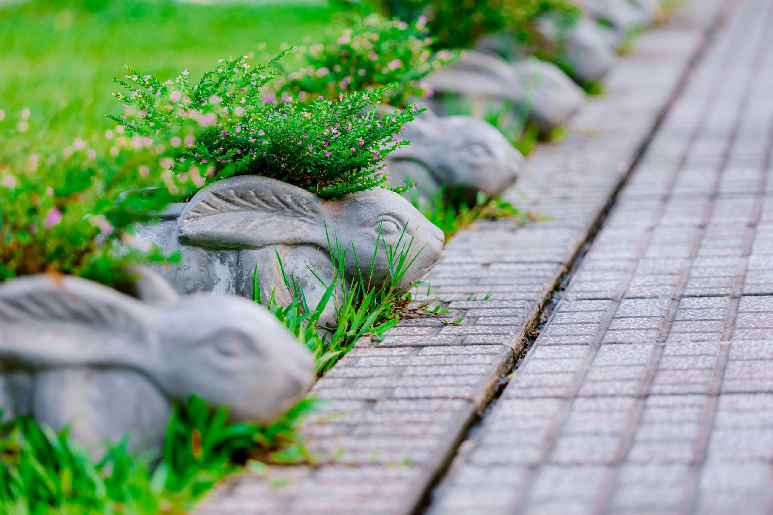 Concrete rabbit planters with flowers line a brick pathway in a vibrant garden setting. Des jardinières en béton en forme de lapin avec des fleurs alignées le long d'une allée en brique dans un cadre de jardin vibrant.