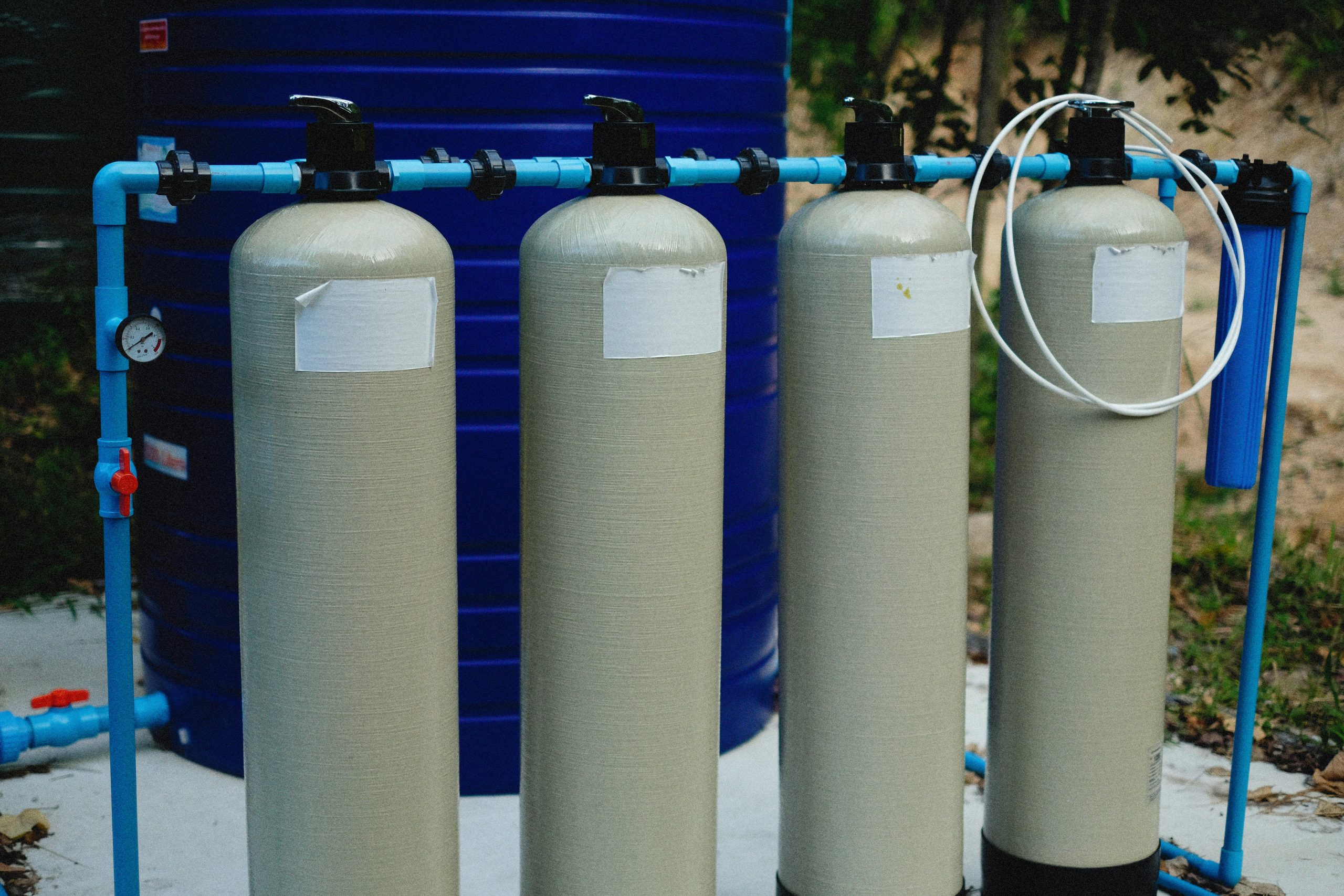 Four beige tanks in an outdoor water treatment setup with blue piping. Quatre réservoirs beige dans une installation de traitement de l'eau extérieure avec tuyauterie bleue.
