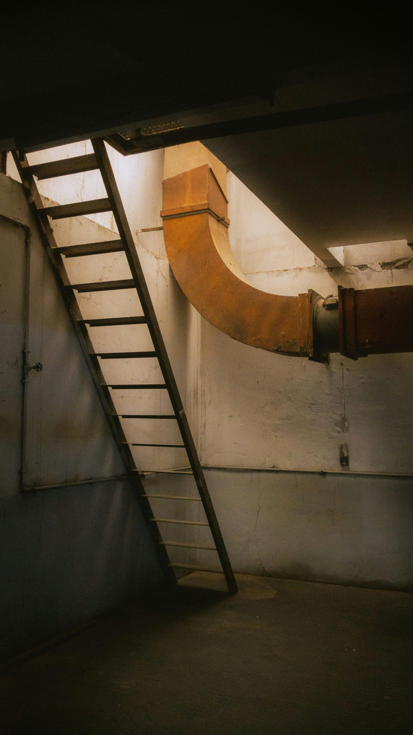 An industrial basement featuring a ladder leading to an upper level and a prominent ventilation pipe. Un sous-sol industriel avec une échelle menant à un niveau supérieur et un tuyau de ventilation prominent.
