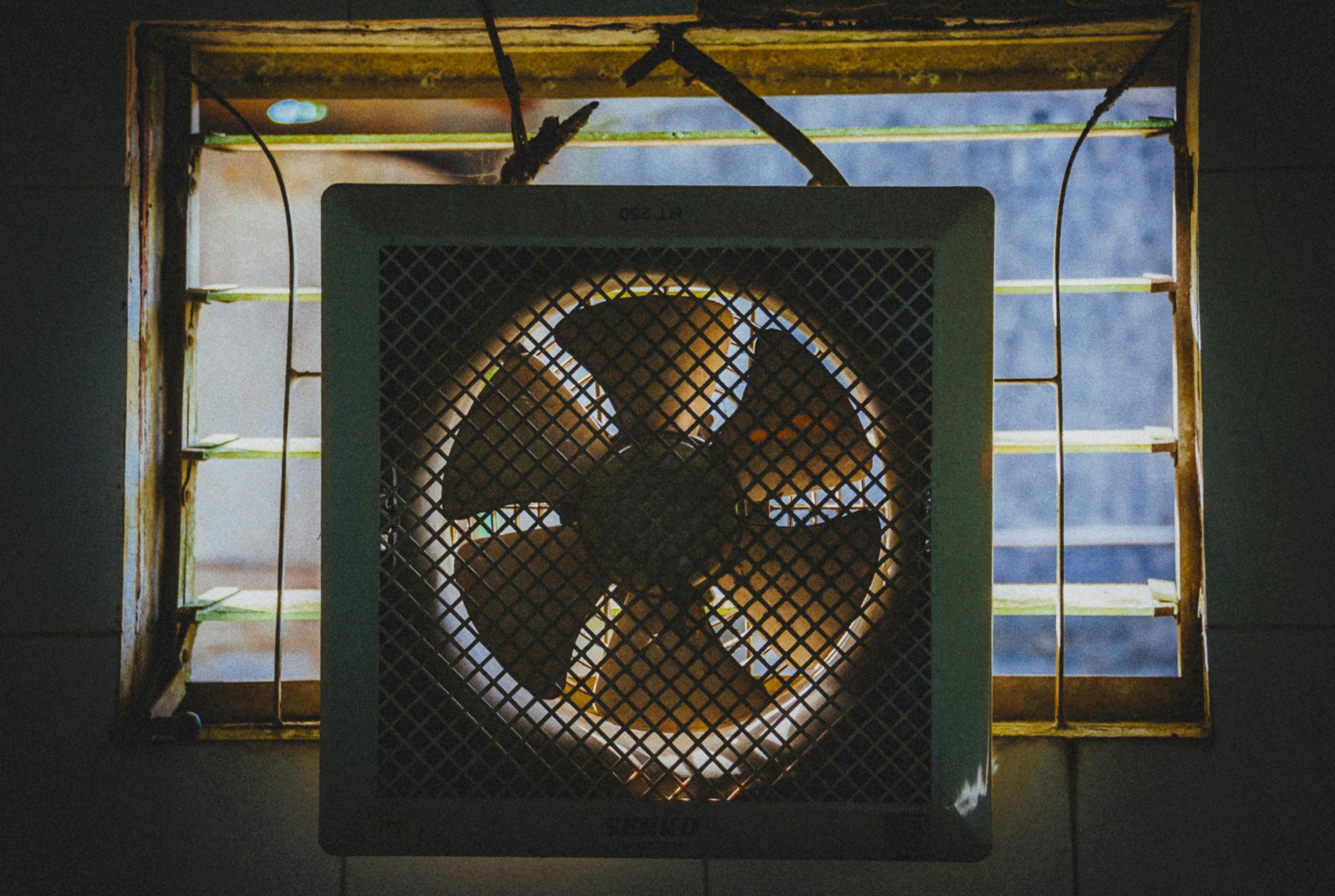 A vintage exhaust fan installed in a rustic window frame, casting dramatic shadows. Un ventilateur d'échappement vintage installé dans un cadre de fenêtre rustique, projetant des ombres dramatiques.