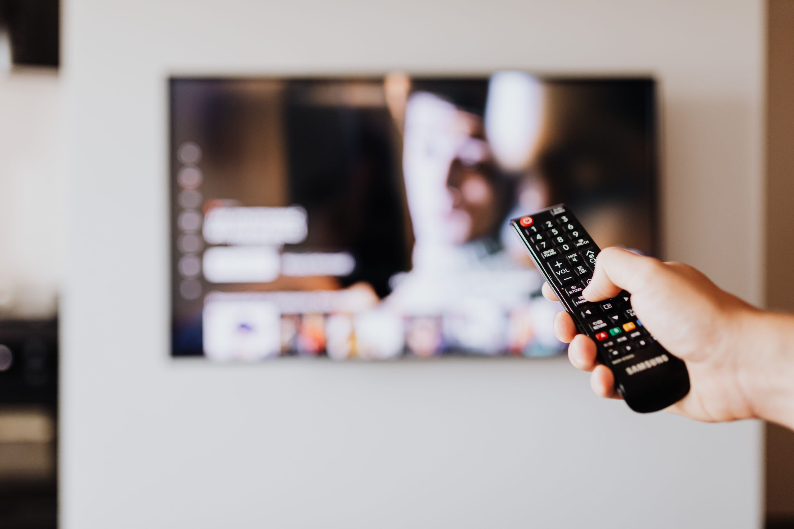 Person using a TV remote control with a blurred television screen in the background. Une personne utilisant une télécommande de télévision avec un écran de télévision flou en arrière-plan.