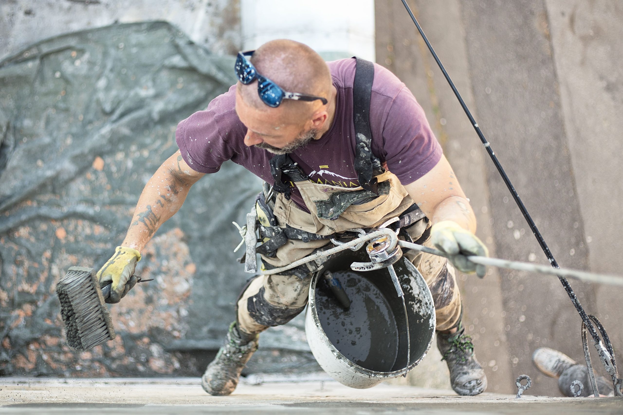 From above of man in safety harness hanging above ground and smoothing wall skim coat with liquid plaster using coarse brush Vue de dessus d'un homme en harnais de sécurité suspendu au-dessus du sol et lissant un enduit mural avec un pinceau grossier en plâtre liquide