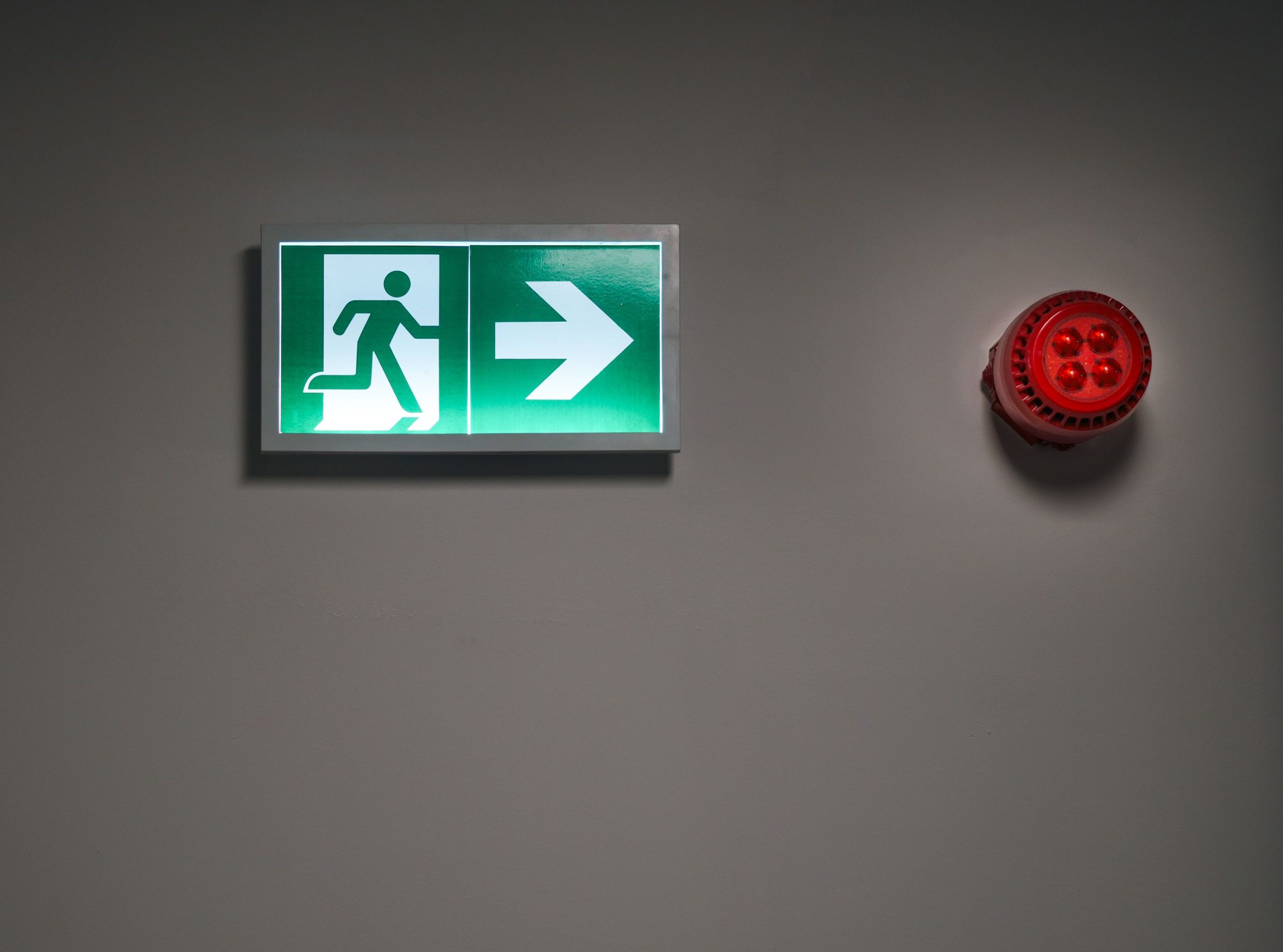 A brightly lit exit sign and alarm in an indoor corridor, indicating emergency escape route. Un panneau de sortie brillamment éclairé et une alarme dans un couloir intérieur, indiquant une route d'échappement d'urgence.