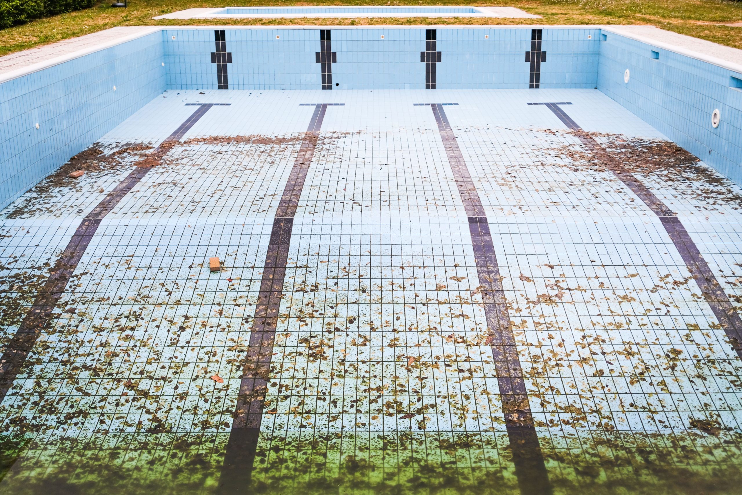 High angle view of an empty, abandoned swimming pool with leaves scattered on the tiled surface. Vue de haut d'une piscine abandonnée et vide avec des feuilles dispersées sur la surface carrelée.