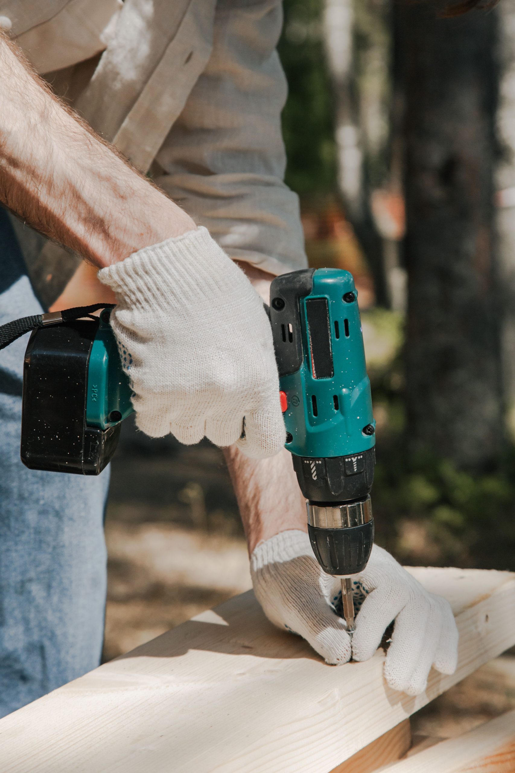 A construction worker using a cordless drill on a wooden panel outdoors, wearing protective gloves. Un ouvrier du bâtiment utilisant une perceuse sans fil sur un panneau en bois à l'extérieur, portant des gants de protection.