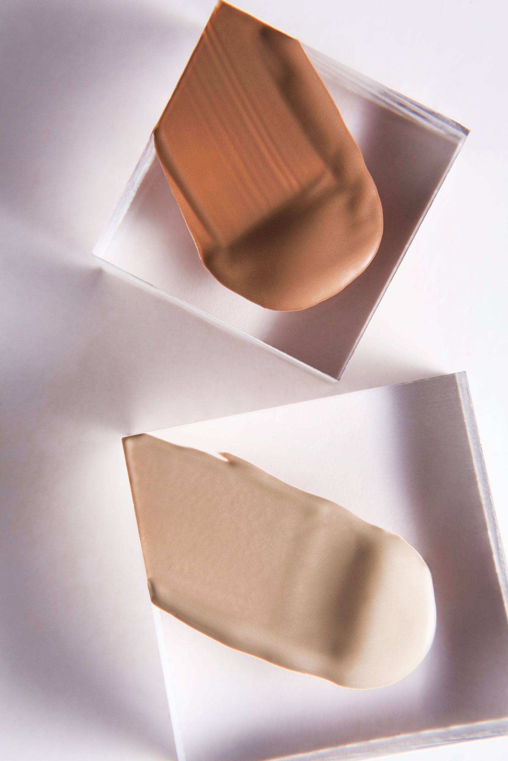 Flat lay image of two cosmetic foundation swatches on a white background, showcasing texture and color. Image en aplat de deux nuances de fond de teint cosmétique sur un fond blanc, mettant en évidence la texture et la couleur.